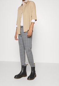 Uomo che indossa una camicia button-up beige a maniche corte sopra una maglietta bianca a maniche lunghe, pantaloni grigi a scacchi e stivali Chelsea neri in pelle.