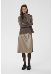 Un pull en tricot marron avec un col haut, associé à une jupe en satin beige, des collants noirs et des chaussures noires polies. Textures lisses et coupe superposée.