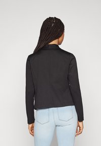Svart tilpasset jakke med krage, lange ermer, og en jevn tekstur; kombinert med lyseblå jeans, som viser en moderne silhuett.