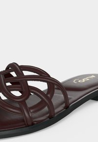 Gros plan d'une sandale plate marron foncé avec des lanières en cuir entrelacées et une semelle noire sur un fond clair.