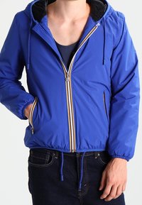 Blaue Kapuzenjacke mit Reißverschluss vorne, weißen und orangen Streifenakzenten, elastischen Bündchen und Kordelzug am Saum. Der Stoff wirkt glatt und leicht.