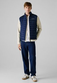 Navy puffer gilet met een verticale gewatteerde stijl, gecombineerd met een lichtgekleurde shirt, blauwe jeans en casual sneakers met beige accenten.
