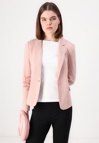 Světle růžový blazer s jedním knoflíkem, klopami a měkkou texturou, v kombinaci s bílým topem a černými kalhotami. Drží růžovou kabelku.