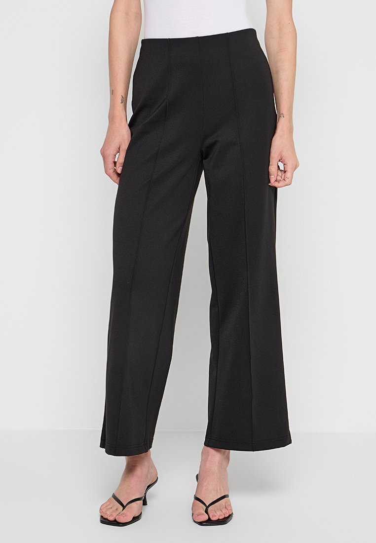 Czarne, szerokie spodnie wykonane z gładkiego, lekkiego materiału, z plisami z przodu i wysokim stanem, w połączeniu z sandałami na cienkich paskach.