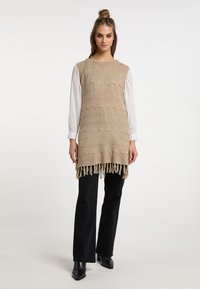 Tunika beige lavorata a maglia con motivi orizzontali e orlo con frange. Indossata sopra una camicia bianca e abbinata a pantaloni neri a gamba larga e stivaletti.