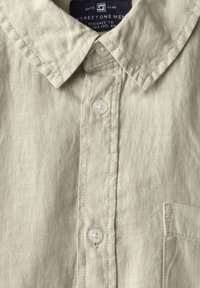 Camisa de lino beige claro con cuello puntiagudo, cierre de botones blancos y un bolsillo en el pecho con sutiles detalles de costura.