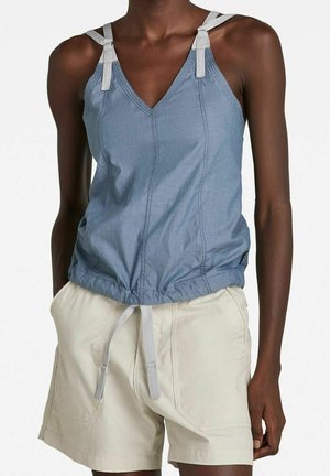 Kvinde iført blå ærmeløs V-hals top med justerbare grå stropper og beige shorts med sidelommer, stående mod en hvid baggrund.