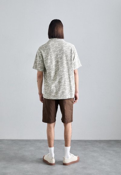 Korte mouwen, lichtgrijze geblokte shirt met bruine shorts. Witte sokken en beige schoenen met bruine zolen. Achteraanzicht tegen een grijze achtergrond.