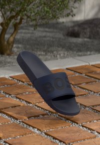 Navy blue slide sandal with "BOSS" lettering floating above wooden square tiles and small white stones outdoors.