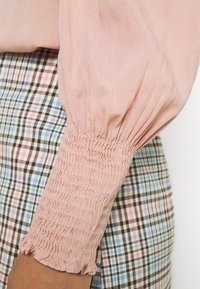 Blouse rose à manches froncées en tissu lisse, associée à une jupe à carreaux présentant des tons bleus, roses et bruns. Détail de tissu texturé visible.