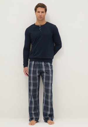 Hombre de pie descalzo con camisa henley de manga larga azul marino y pantalones de pijama a cuadros azul marino y blanco sobre un fondo liso.