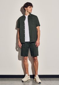 Kortärmad mörkgrön linneskjorta med en framlappficka, matchad med shorts i samma färg och beigea sportskor mot en neutral bakgrund.