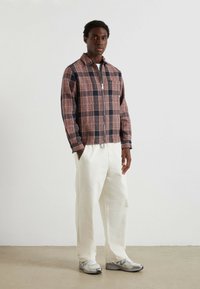 Mężczyzna stoi w różowo-czarnej kratkowanej kurtce na zamek, białych szerokich spodniach i srebrnych butach do biegania na tle białej ściany.