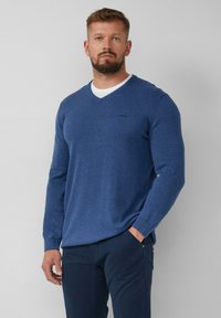 Blauer V-Ausschnitt-Pullover aus Strickstoff, mit langen Ärmeln und einem kleinen Logo auf der Brust, kombiniert mit dunklen Hosen.
