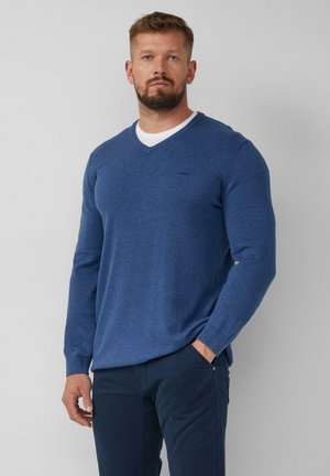 Pull bleu en v en tissu tricoté, avec des manches longues et un petit logo sur la poitrine, associé à un pantalon sombre.