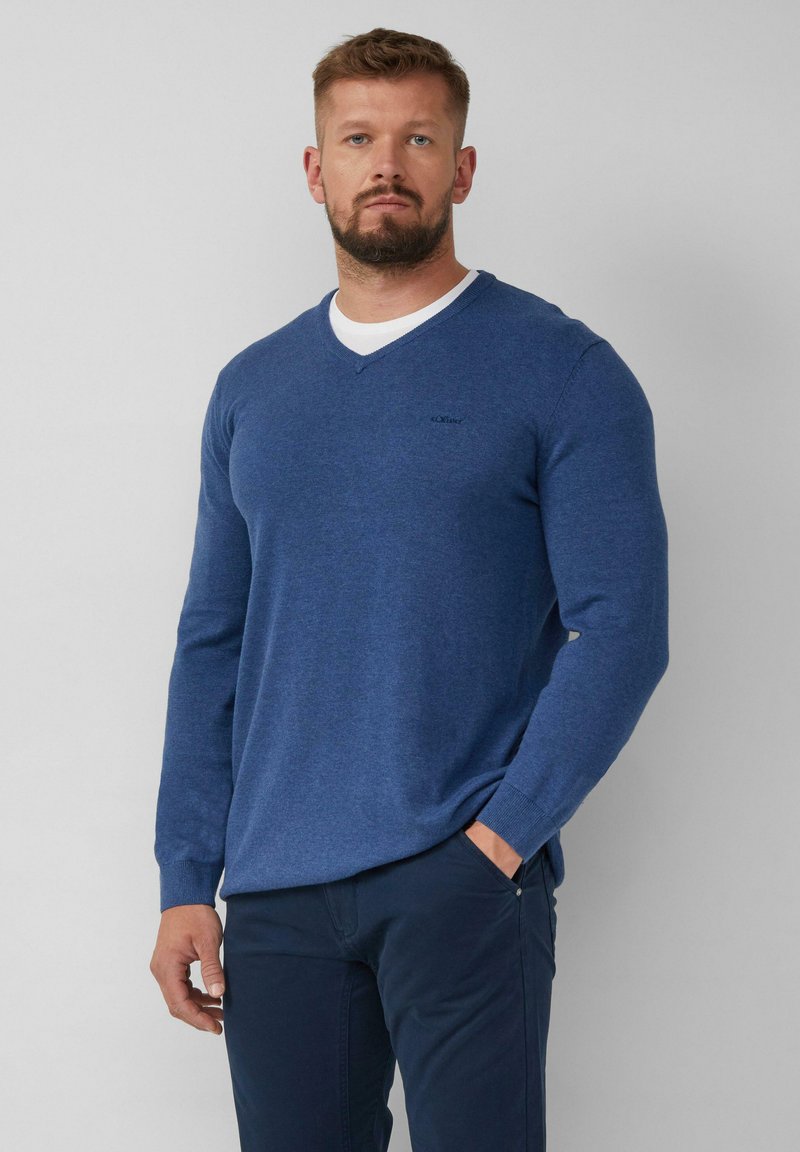 Blauer V-Ausschnitt-Pullover aus Strickstoff, mit langen Ärmeln und einem kleinen Logo auf der Brust, kombiniert mit dunklen Hosen.