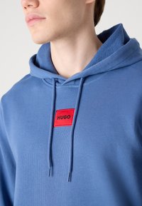 Blå hettegenser laget av mykt stoff, med et rødt rektangulært logo-patch med teksten "HUGO" i midten. Snorene er til stede.