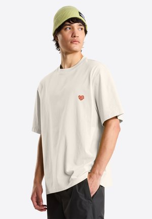 Ung mand iført en lysebeige North Face T-shirt, sorte bukser med den ene hånd i lommen og en lysegrøn strikket hue, der kigger til siden.