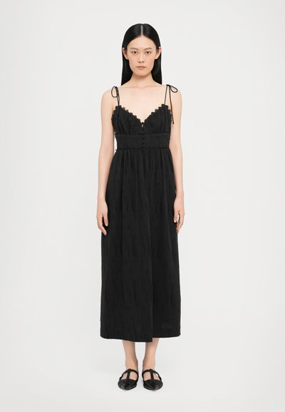 Robe maxi noire avec bord en dentelle et bretelles spaghettis réglables, comportant une fermeture à boutons et un tissu texturé. Le mannequin porte des chaussures noires plates.