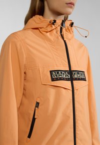 Lichte oranje jas met een rits, een verstelbare capuchon en een borstzak met een zwart-wit logo. Geststructuurde stof.