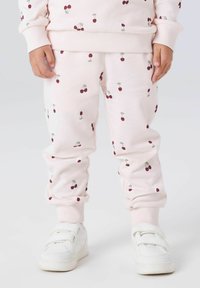 Dziecko w jasnoróżowych spodniach z nadrukiem czereśni i białych trampkach na rzep, stojące na jednolitym tle.