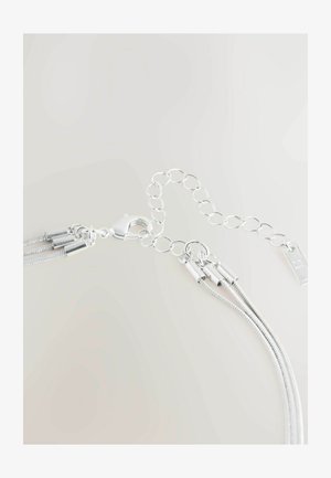 Sudraba ķēde ar omāriņa sprādzi, kas satur trīs teksturētas sloksnes un apaļus gredzenus. Pievienota maza taisnstūra birka uz ķēdes.