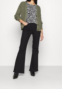 Olivgrön stickad cardigan med ribbad textur och knappar, matchad med svarta utsvängda jeans och en top med svart och vit leopardmönster. Svarta stövlar.