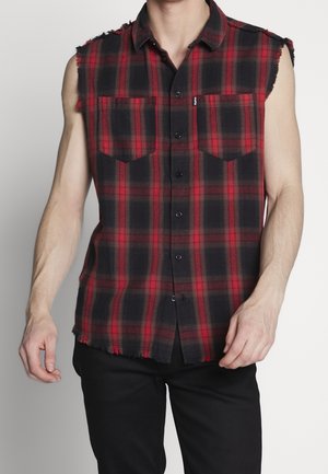 Męska koszula bez rękawów w czerwono-czarną kratę, zapinana na guziki, z postrzępionymi brzegami, noszona z czarnymi spodniami na jednolitym tle.