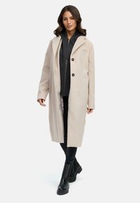Lange beige jas van een wolmix, met een reverskraag, twee voorzakken en knoopsluiting over een donkere rits-hoodie en zwarte broek.