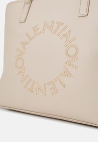 Beige läder tote-väska med präglade guldfärgade nitar arrangerade i ett cirkulärt mönster, med text i versaler.