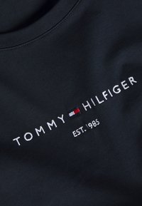 Zbliżenie na granatową tkaninę z białym haftowanym napisem "Tommy Hilfiger", małym logo w kolorach czerwonym, białym i niebieskim, oraz "EST. 1985" poniżej.