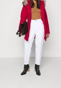 Czerwona parka z futrzanym kapturem, brązowy prążkowany top, białe spodnie i czarne botki na obcasie. Model z zamkiem błyskawicznym i zapięciem na guziki, o eleganckim kroju i teksturowanych tkaninach.