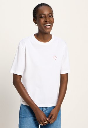 Lächelnde Frau mit geflochtenem Haar trägt ein weißes T-Shirt mit einem kleinen roten Herz auf der Brust, blaue Jeans und goldene Ringe an ihren Fingern.