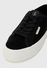 Black corduroy sneaker with textured pattern, black laces, and white rubber sole. Features a "Calvin Klein" logo tag on the side.