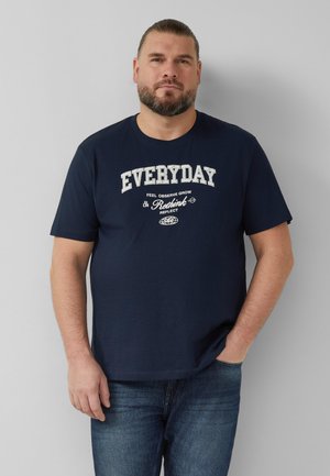 Mężczyzna z brodą, ubrany w granatowy T-shirt z napisem „EVERYDAY” i innym nadrukiem, stoi z jedną ręką w kieszeni jeansów na tle szarej ściany.