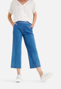 Personne portant un jean bleu à taille large et courte avec une ceinture élastique, un blouse blanche à col en V et manches courtes, des baskets blanches, posant avec les mains dans les poches.