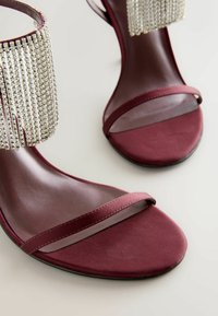 Burgunderfarbene Absatzsandalen mit einem eleganten Design, ausgestattet mit einem dünnen Riemen und einem knöchelhohen Bund, der mit Kristallen verziert ist und vertikale Linien aufweist.