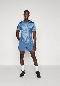 Mężczyzna stoi z rękami w kieszeniach, ubrany w niebieską sportową koszulkę w stylu tie-dye, dopasowane szorty, białe skarpetki i czarne buty do biegania.