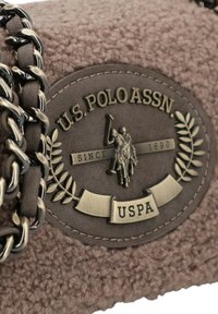 Materiale marrone e soffice presenta un'emblema in bronzo della U.S. Polo Assn. con una scena di polo, circondata da foglie di alloro e accenti decorativi a catena.