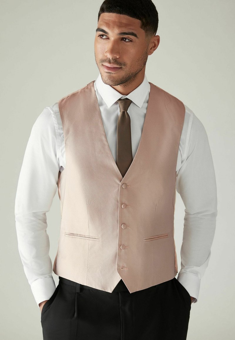 Gilet texturé rose clair avec un décolleté en V profond, cinq boutons et deux poches avant, porté sur une chemise blanche et une cravate marron.
