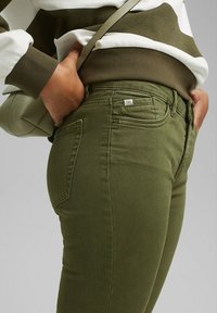 Personne portant un jean vert ajusté et un sweat-shirt rayé vert et blanc, les mains dans les poches arrière et une sangle de sac à main verte.