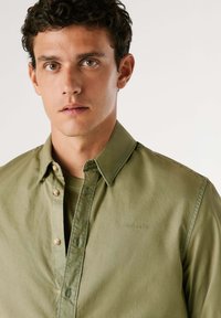 Camicia button-up verde oliva con una texture morbida, caratterizzata da un colletto aperto e un sottile ricamo del logo sul petto. Finitura leggermente stropicciata.