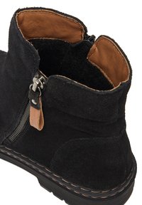 Botín de ante negro con forro de cuero color canela, cremallera lateral y costuras en contraste a lo largo de la suela. Textura suave y diseño redondeado.