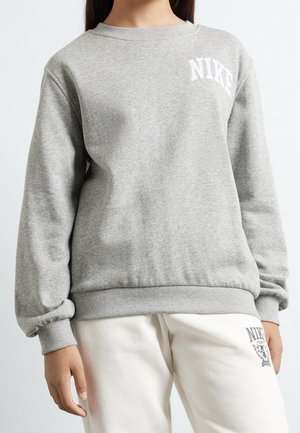 Sweat-shirt gris avec col rond, poignets et ourlet côtelés. Présente un logo "NIKE" blanc sur le côté gauche de la poitrine. Associé à un pantalon de couleur claire.