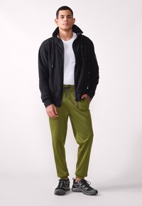 Chaqueta negra con cremallera y cuello alto, camiseta blanca, pantalones verdes con cintura elástica y zapatillas deportivas negras con detalles.