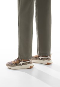 Sneakers multicolori con un motivo beige, marrone e bianco, dotati di una suola imbottita e lacci visibili, abbinati a pantaloni verde oliva.