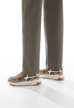 Mehrfarbige Sneaker mit einem beigen, braunen und weißen Muster, ausgestattet mit einer gepolsterten Sohle und sichtbaren Schnürsenkeln, kombiniert mit olivgrünen Hosen.