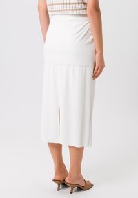 Witte mid-length rok met een split aan de achterkant, gemaakt van een gladde stof. Gecombineerd met bruine hoge sandalen, wat een gestroomlijnde silhouet toont.