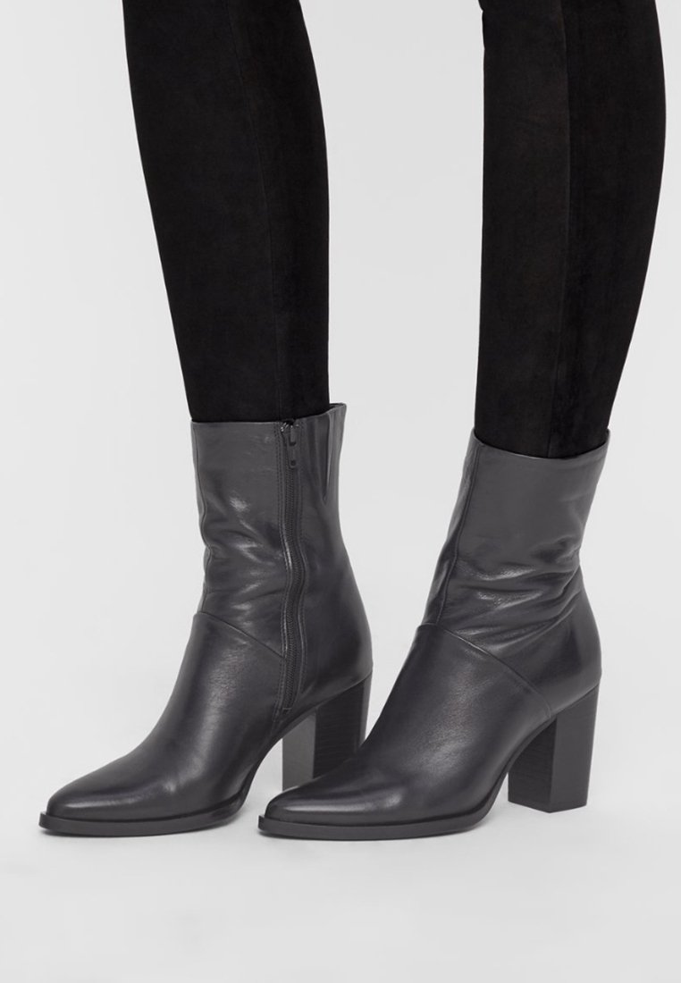 Bottes chevillées en cuir noir avec bouts pointus, fermetures éclair latérales et talons carrés, portées avec un pantalon noir.