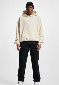 Sweat à capuche beige avec une poche frontale, associé à un pantalon cargo noir. Le modèle porte un bonnet noir et des baskets blanches.